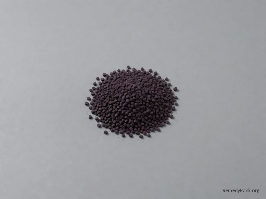 A small heap of dark purple potassium permanganate crystals in a plain glass dish, photographed plainly on a neutral grey background.